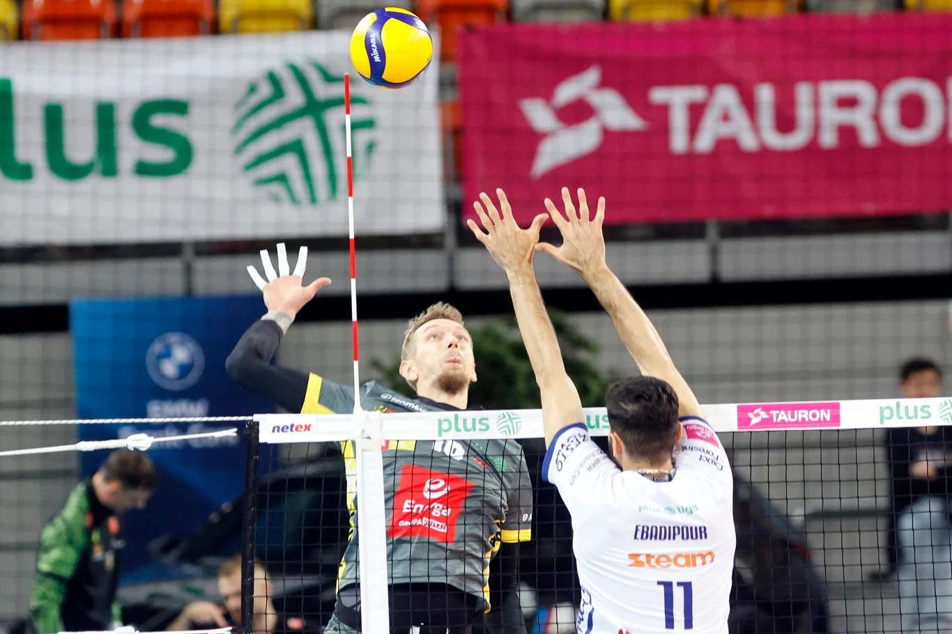 Dwóch siatkarzy w akcji przy siatce, jeden wykonuje atak, drugi próbuje blokować po drugiej stronie boiska. Piłka znajduje się nad siatką, a w tle widoczne są trybuny oraz banery reklamowe.