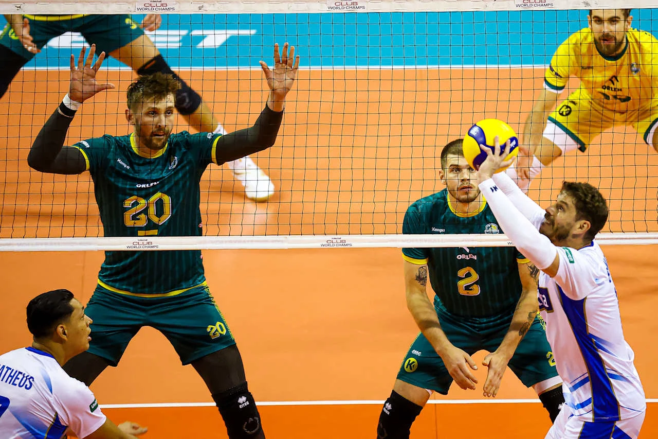 Sportowa rywalizacja na siatkarskim boisku, kilku zawodników w barwach dwóch drużyn w dynamicznej akcji przy siatce, zawodnik w białym stroju podbija piłkę, a przeciwnicy w zielono-żółtych strojach przygotowują się do bloku.