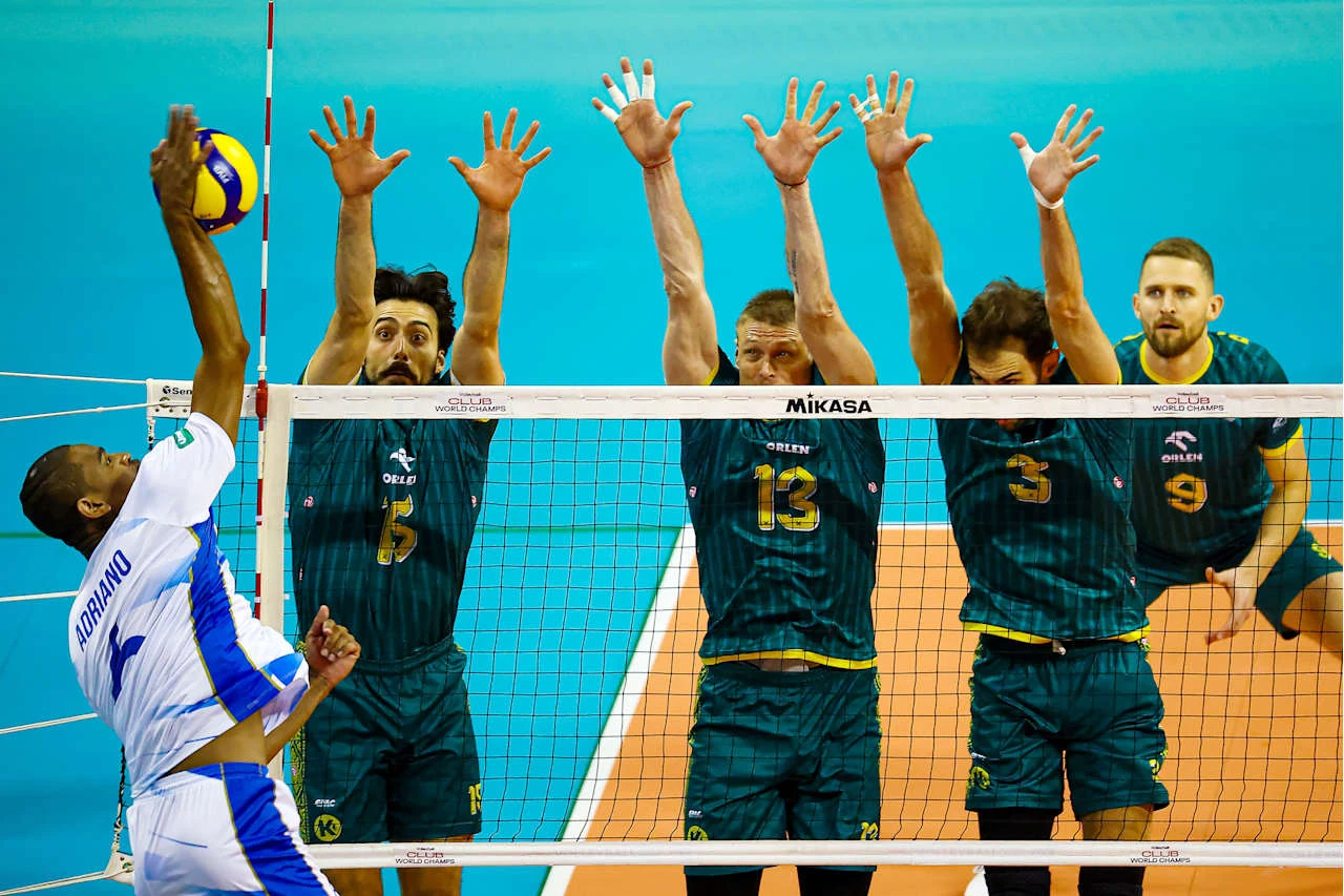 Siatkarz drużyny w białym stroju atakuje piłką ponad siatką, podczas gdy trzech zawodników drużyny przeciwnej w ciemnozielonych strojach próbuje zablokować piłkę, unosząc ręce do góry. W tle widoczny czwarty zawodnik obserwujący sytuację.