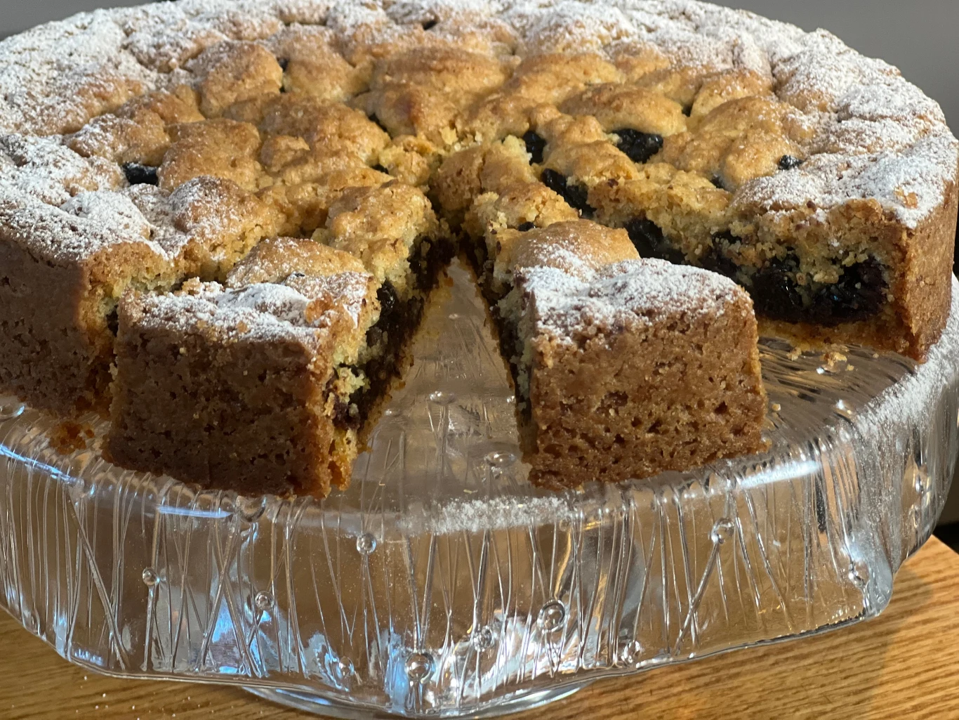 Ciasto drożdżowe z owocowym nadzieniem i kruszonką, posypane cukrem pudrem, ułożone na szklanej paterze, z wyciętym kawałkiem eksponującym wnętrze wypieku.