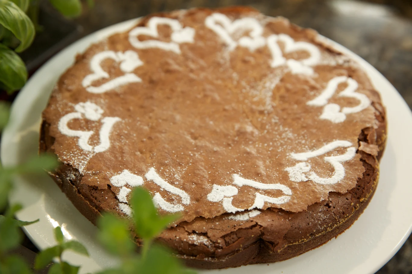 Okrągłe, brązowe ciasto z dekoracją z cukru pudru w kształcie serduszek położone na białym talerzu, z zieloną rośliną w tle.