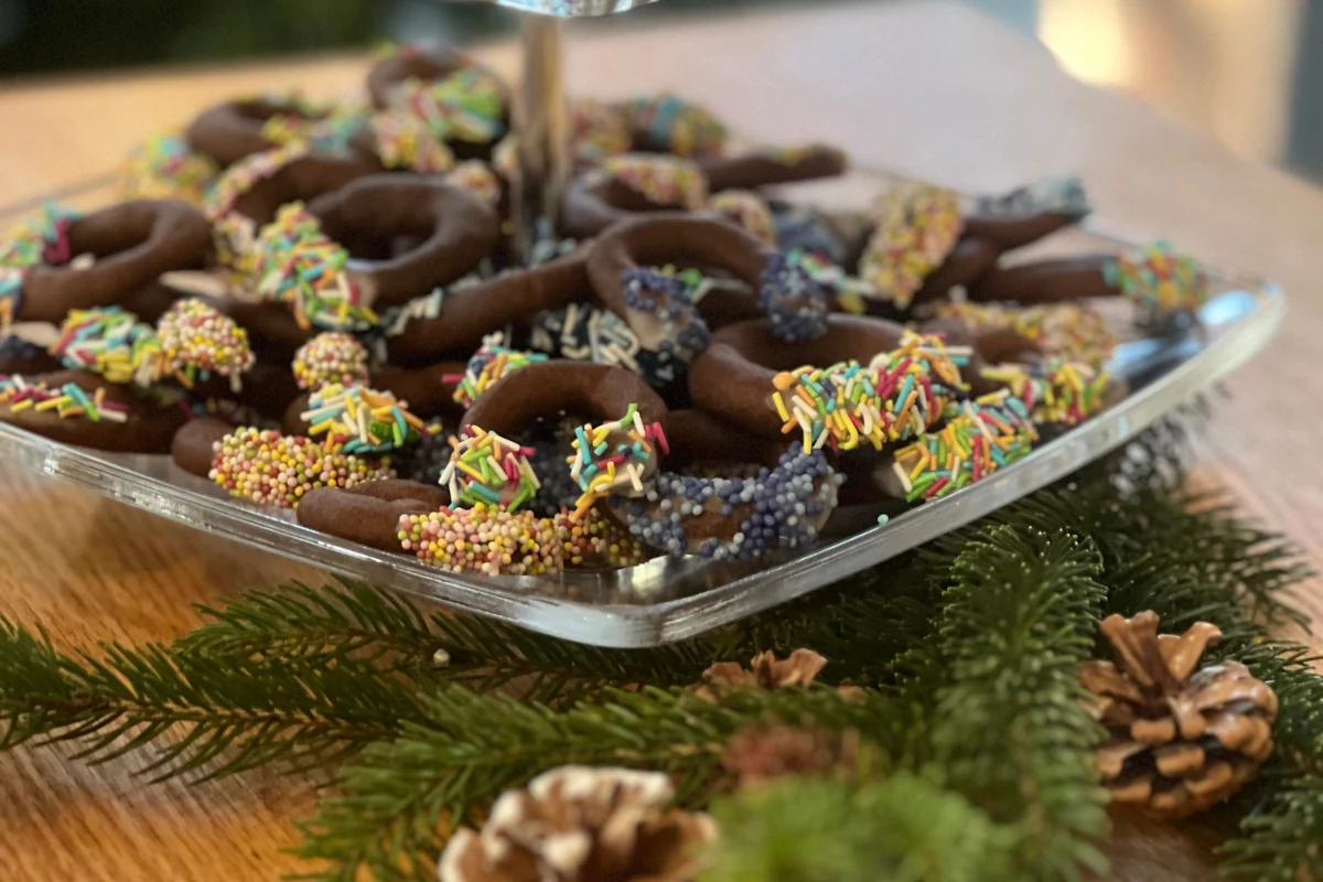 Półmisek z czekoladowymi ciasteczkami ozdobionymi kolorowymi posypkami, otoczony gałązkami świerkowymi i szyszkami na drewnianym stole.