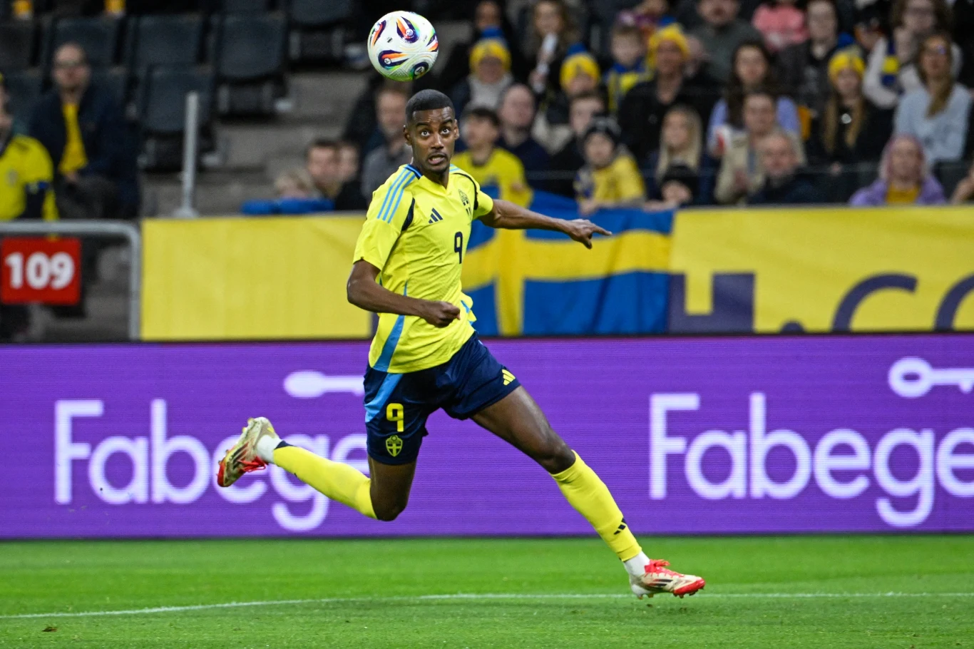 Piłkarz w żółtej koszulce i granatowych spodenkach w trakcie meczu, biegnący za piłką na stadionie, tłum kibiców w tle, dominują żółto-niebieskie barwy.