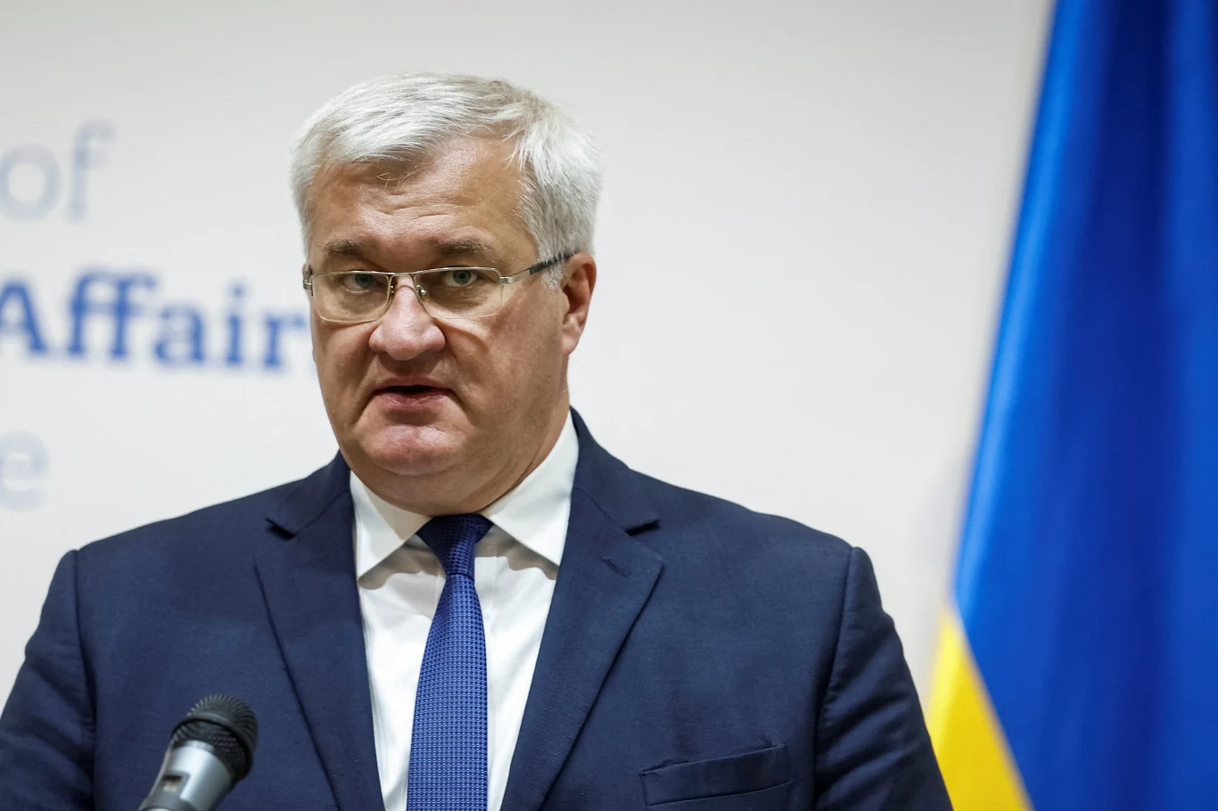 Mężczyzna w garniturze i okularach stoi przy mównicy, w tle fragment flagi Ukrainy i rozmazany napis angielski na ścianie.