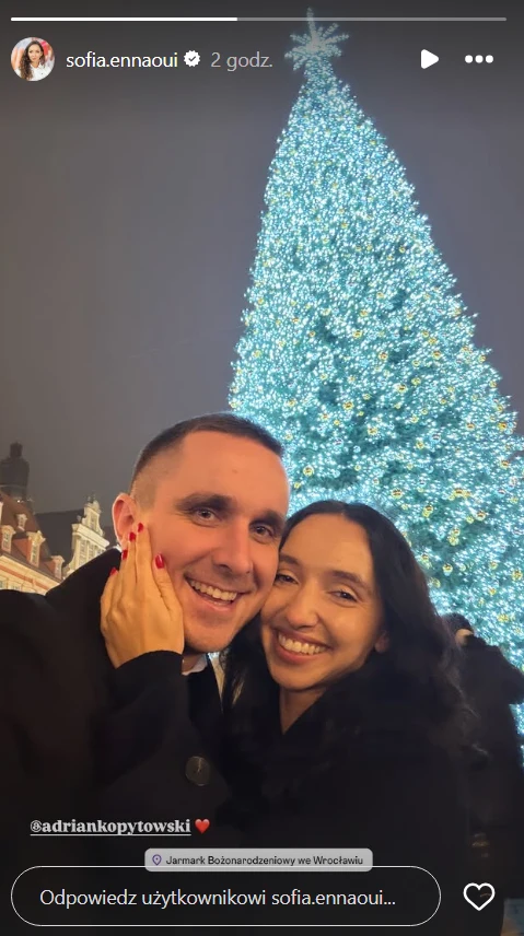 Uśmiechnięta para obejmuje się na tle wysokiej, jasno oświetlonej choinki bożonarodzeniowej na miejskim rynku. Ich twarze wyrażają szczęście i bliskość, wokół panuje świąteczna atmosfera nocą.