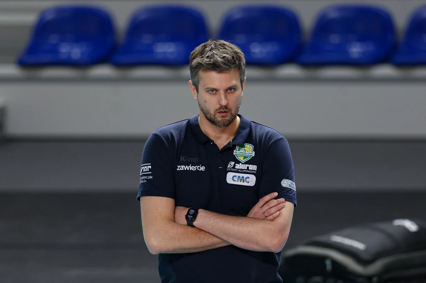 Mężczyzna w sportowej koszulce z logo klubu, stojący z założonymi rękami na tle pustych niebieskich krzeseł.