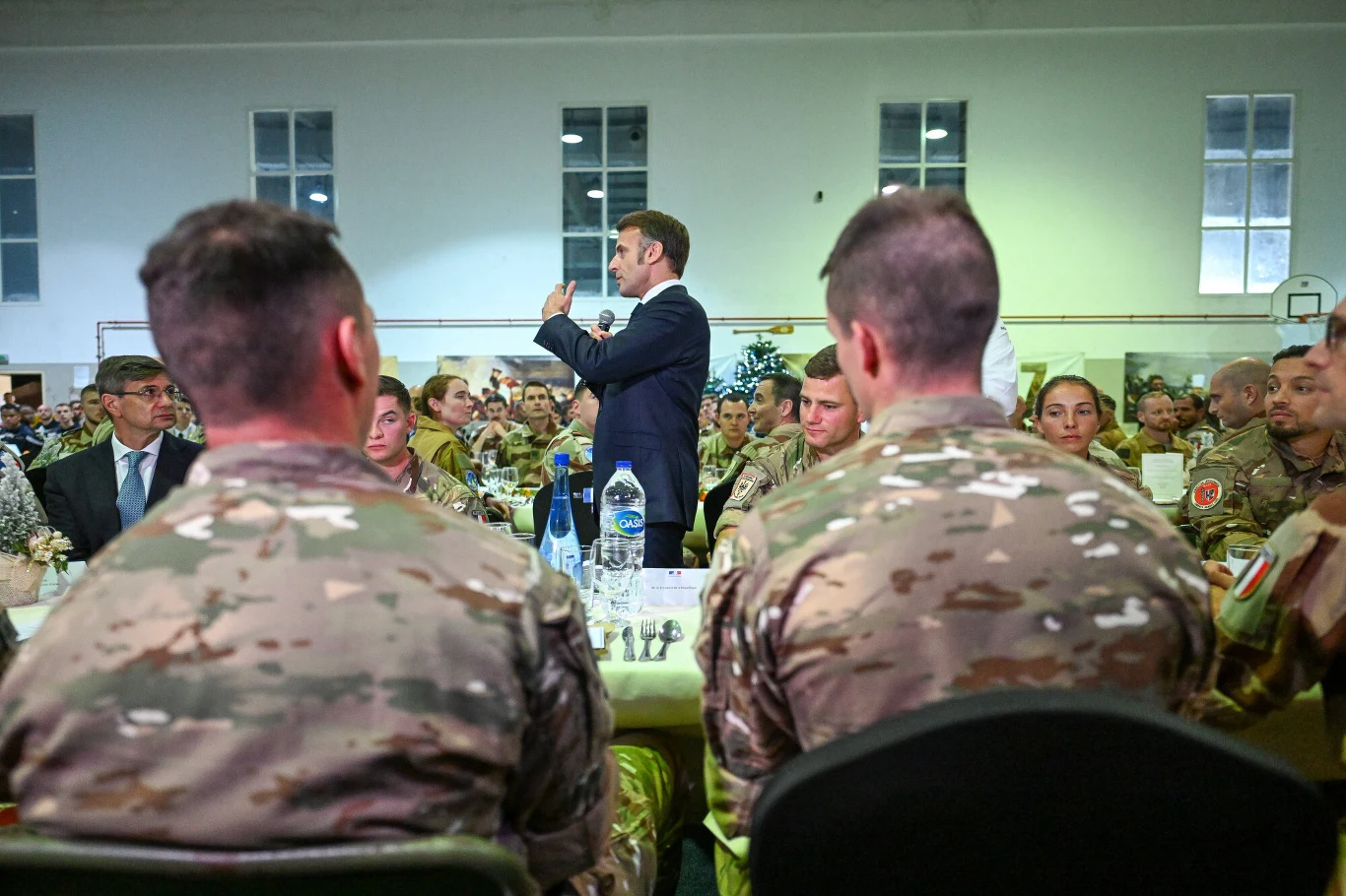 Oficjalne spotkanie wojskowych z cywilnym przywódcą w dużej, dobrze oświetlonej hali. Na pierwszym planie żołnierze w mundurach siedzący przy stole, za nimi liczne osoby w tle. Centralną postacią jest mężczyzna stojący, przemawiający do zebranych.