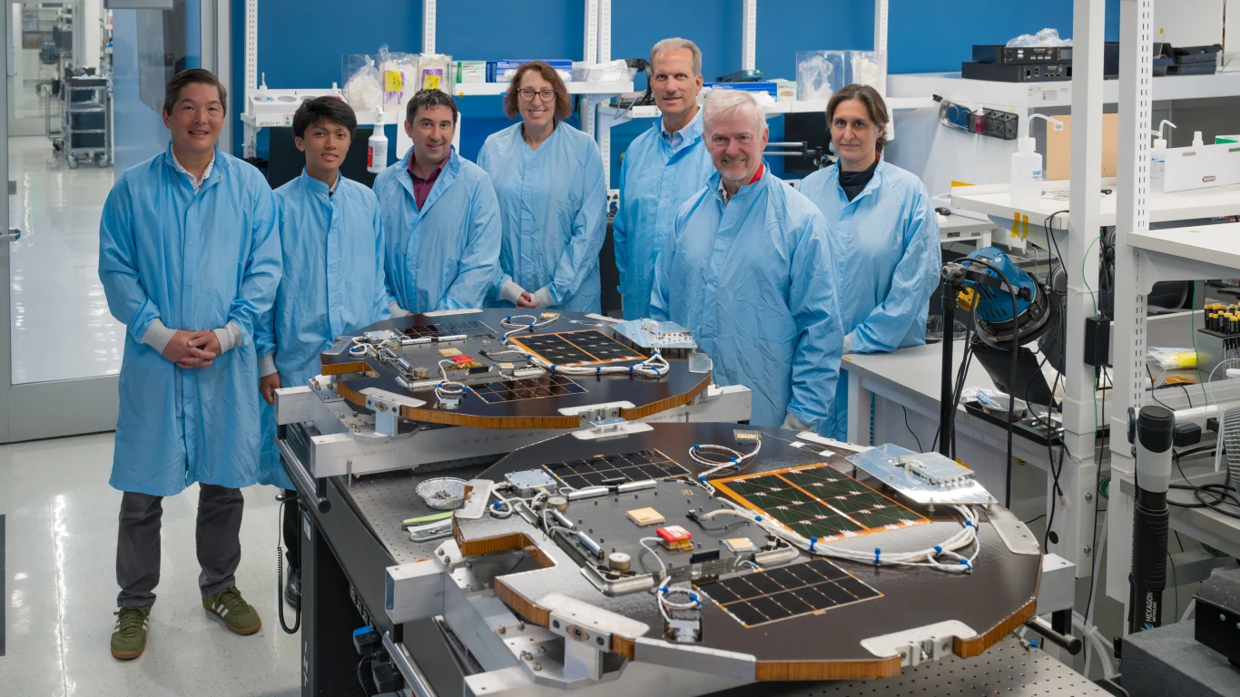 Grupa inżynierów w niebieskich fartuchach laboratoryjnych stoi wokół stołów, na których znajdują się zaawansowane urządzenia elektroniczne i komponenty, w jasnym, dobrze wyposażonym laboratorium.