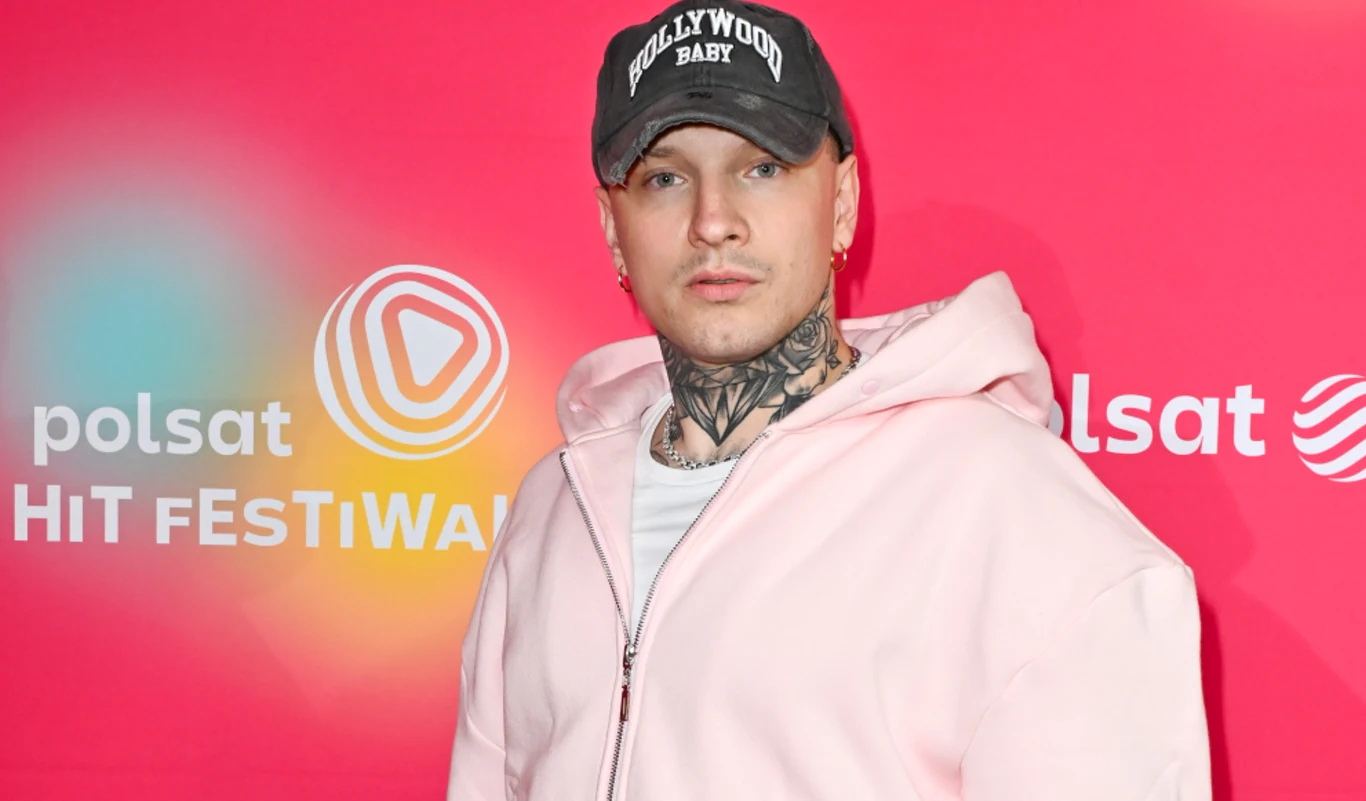 Smolasty na Sylwestrowej Mocy Przebojów 2025 Mężczyzna w jasnoróżowej bluzie z odkrytym tatuażem na szyi i czapce z daszkiem z napisem, stojący na tle ścianki z logo Polsat Hit Festiwal.