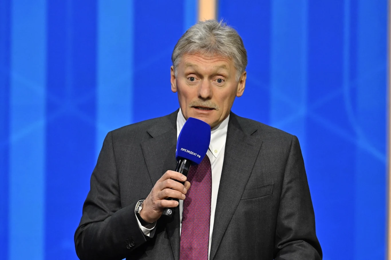 Kreml reaguje na doniesienia agencji Reutera. Dmitrij Pieskow zabrał głos Mężczyzna w szarym garniturze, jasnej koszuli i bordowym krawacie trzyma niebieski mikrofon i przemawia na tle błękitnej, graficznej ściany.