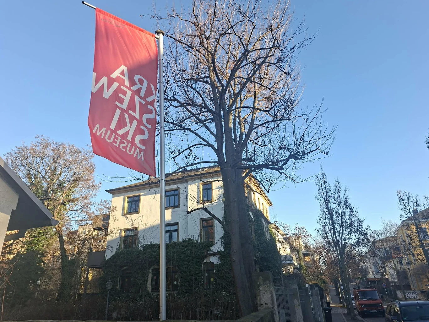 Czerwony baner z napisem „Muzeum Sztuki Współczesnej” powiewa na maszcie przy ulicy wśród budynków mieszkalnych, typowych dla miejskiej zabudowy; w tle drzewa bez liści oraz fragmenty aut zaparkowanych przy drodze.