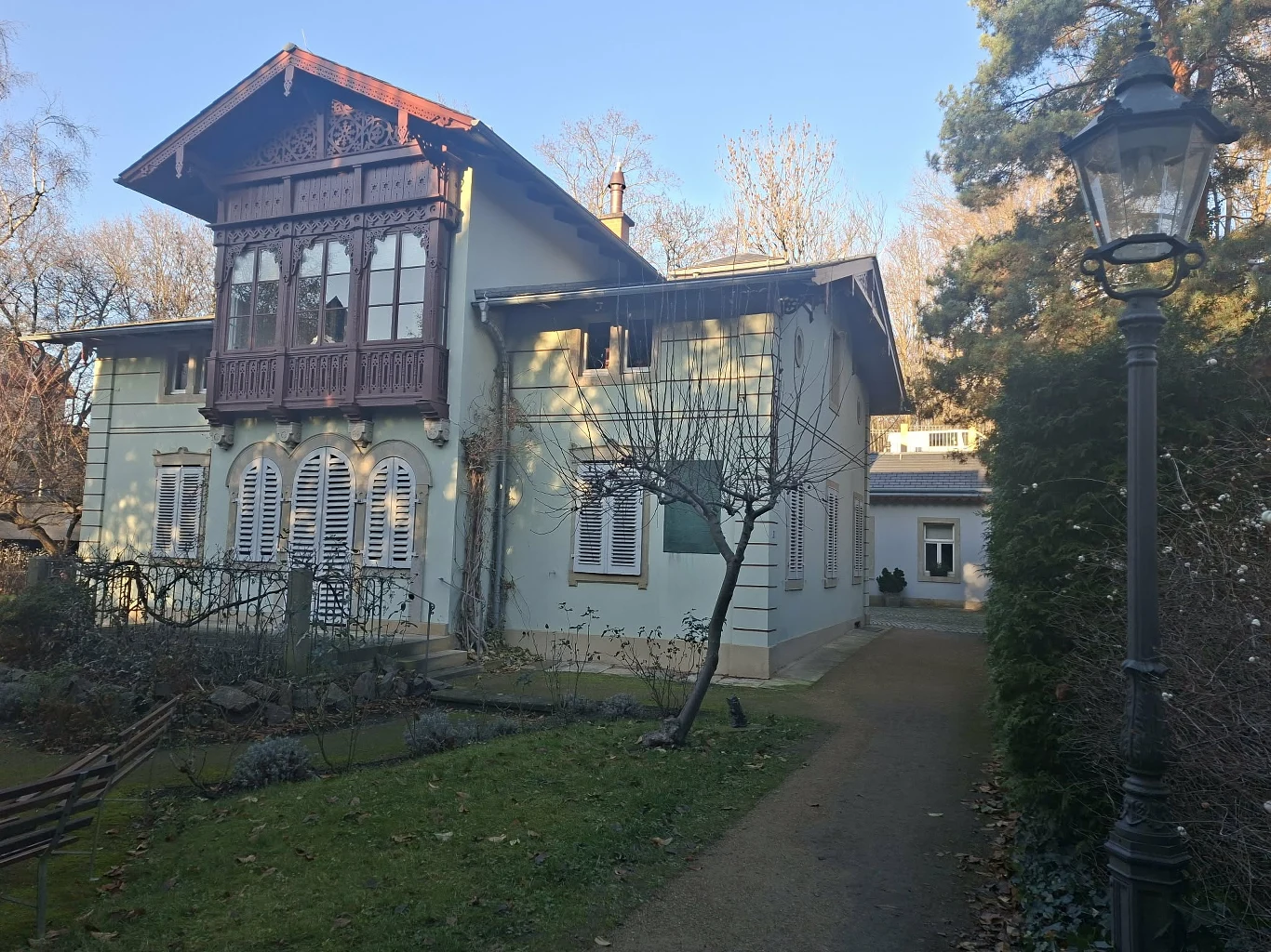 Dwupiętrowy dom w stylu willowym z jasną elewacją i bogato zdobionym balkonem z ciemnego drewna. Przed budynkiem ogród z przyciętymi krzewami oraz alejka prowadząca wzdłuż domu. Po prawej stronie widoczna latarnia ogrodowa.