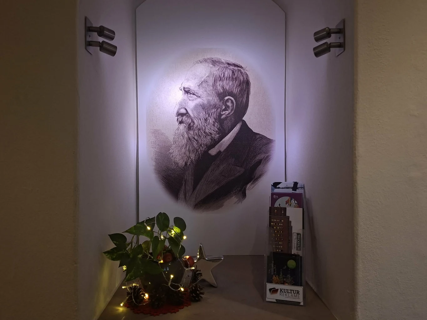 Portret starszego mężczyzny z brodą, ukazany z profilu, zawieszony na ścianie w niszy, poniżej dekoracje w postaci doniczki z rośliną, świątecznego oświetlenia oraz metalowej gwiazdy, obok pamiątkowy zestaw w opakowaniu.