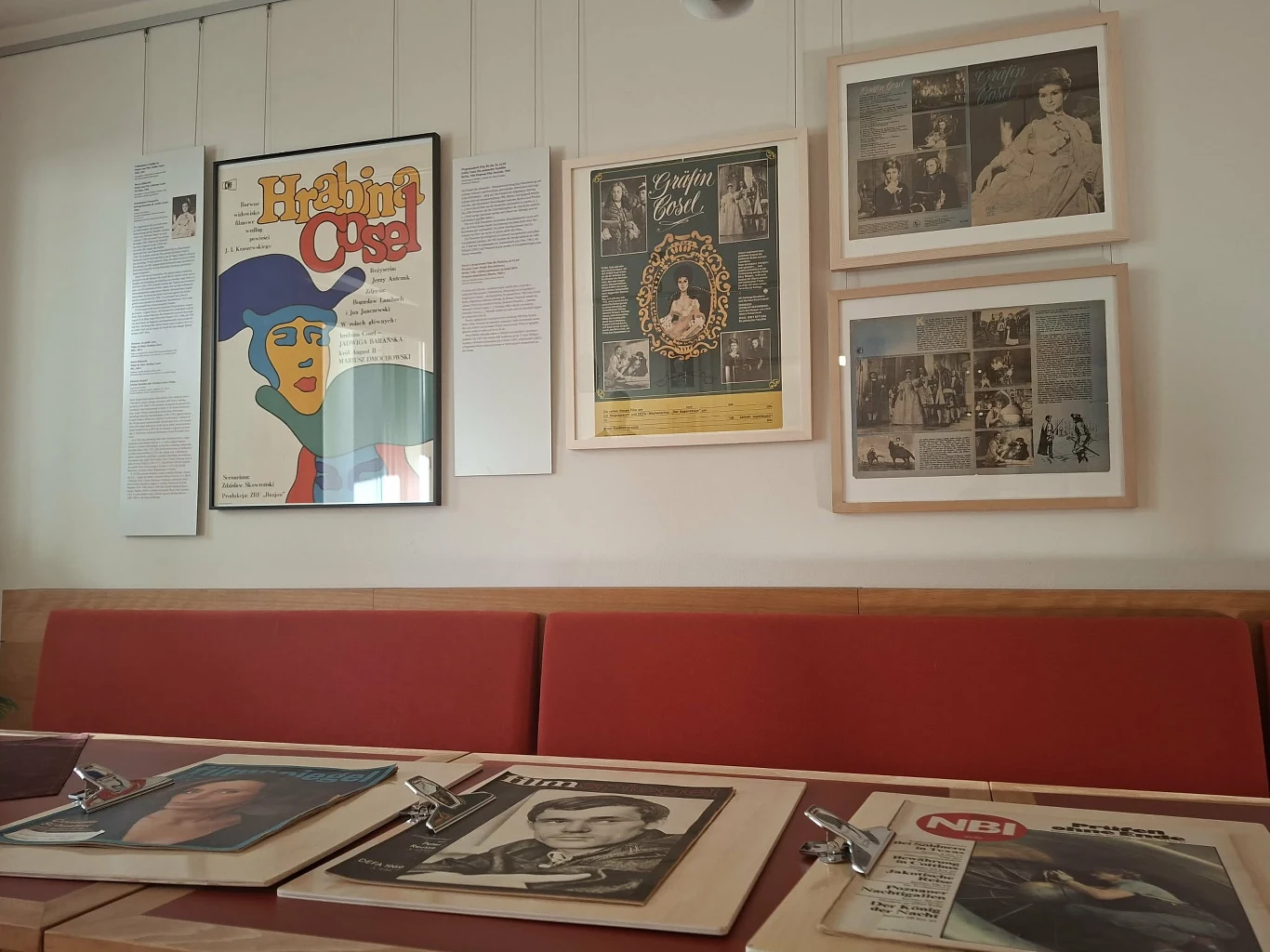 Ściana ozdobiona plakatami i archiwalnymi stronami gazet związanych z kulturą, zdjęcia i grafiki w ramkach, a na pierwszym planie na stole leżą stare czasopisma w plastikowych teczkach z widocznymi okładkami.