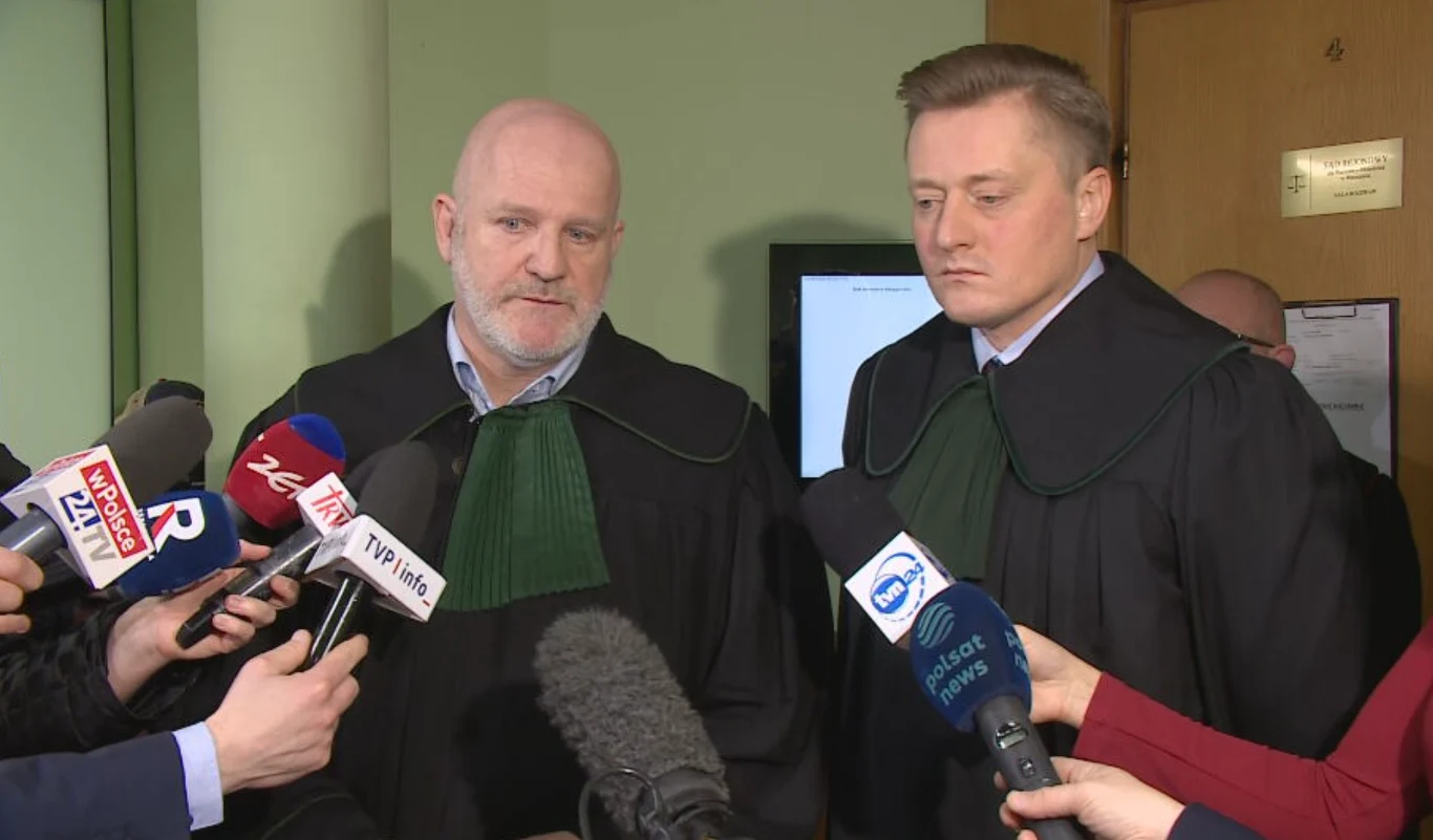 Przedstawiciele prawni byłego szefa MS Zbigniewa Ziobry Dwóch mężczyzn w togach prawniczych udziela wywiadu mediom, mikrofony z logo różnych stacji telewizyjnych skierowane w ich stronę, w tle fragment ściany oraz drzwi.