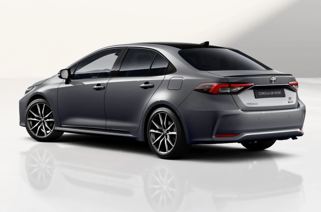 Szary sedan Toyota Corolla prezentowany z tyłu na tle jasnego, neutralnego tła. Nowoczesna sylwetka auta z przyciemnianymi szybami, aluminiowymi felgami oraz charakterystycznym subtelnym spojlerem i wyrazistymi światłami tylnymi.