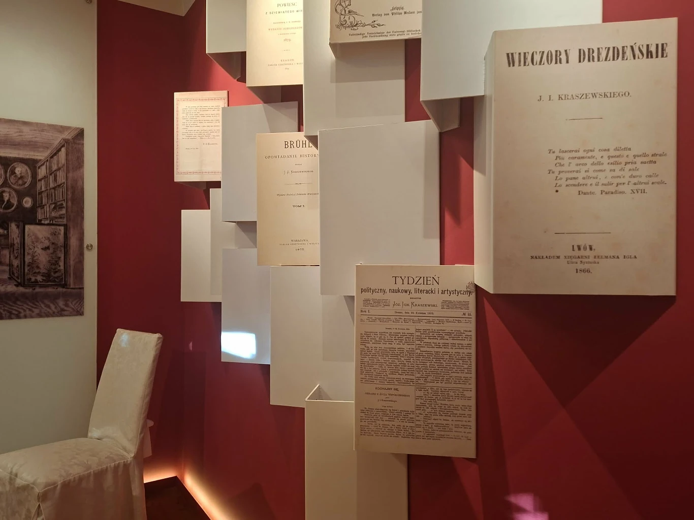 Fragment ekspozycji muzealnej z białym krzesłem oraz ścianą ozdobioną reprodukcjami dawnych dokumentów i druków, z których jeden zatytułowany jest "Wieczory Drezdeńskie".