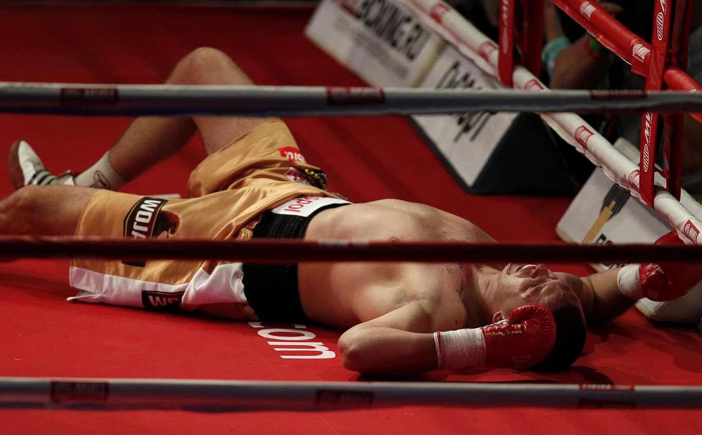 Bokser w złotych spodenkach leży bezwładnie na ringu, tuż przy linach. Jego twarz oraz część ciała zasłonięte są przez ręce w czerwonych rękawicach bokserskich. Widoczne fragmenty czerwonej maty ringu oraz elementy barierek oddzielających ring od widowni.
