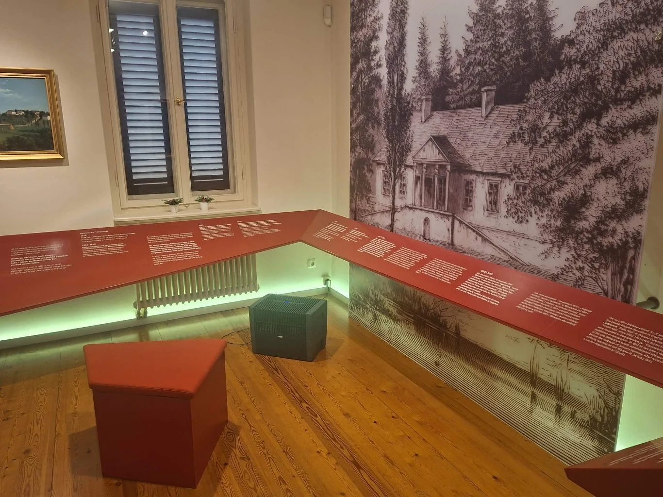 Wnętrze muzealnej sali z ekspozycją, na środku dwa siedziska, wokół drewniana podłoga, na ścianach wystawa z planszami informacyjnymi o czerwonym tle, jedna ściana ozdobiona dużą grafiką przedstawiającą zabytkowy dom otoczony drzewami, w tle okno z zam...