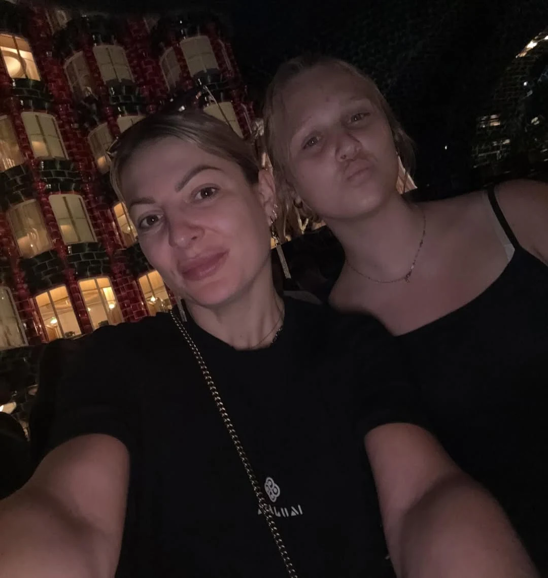 Dwie kobiety pozujące do selfie wieczorem, w tle kolorowo podświetlony budynek z dużymi oknami.