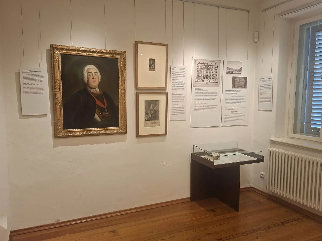 Wnętrze muzeum z eksponatami na ścianie, w tym portretem starszego mężczyzny w ramie oraz kilkoma dokumentami i rycinami; po prawej stronie znajduje się przeszklona gablota z eksponatem oraz okno z żaluzjami.