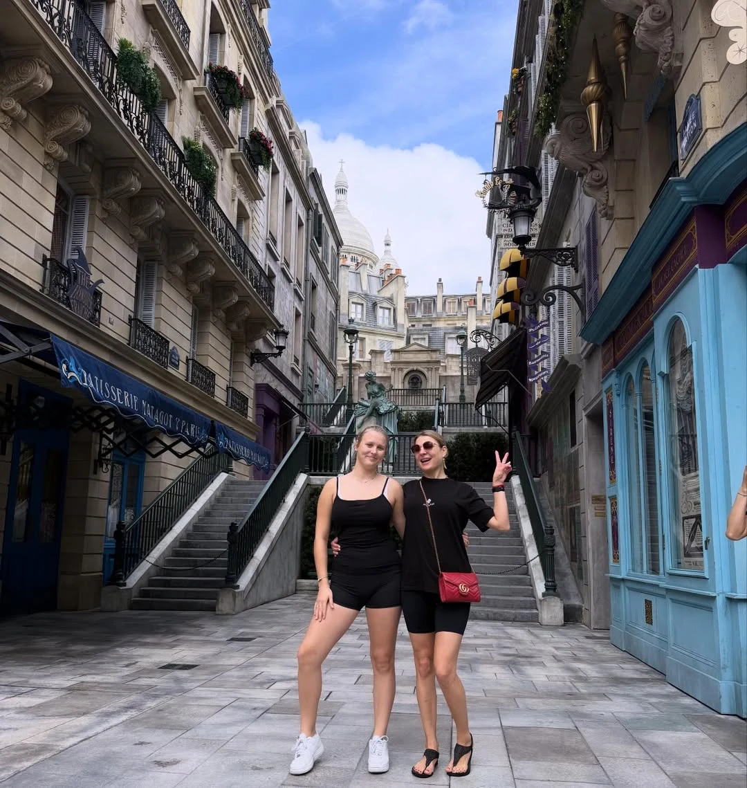 Dwie młode kobiety uśmiechające się i pozujące na tle malowniczej miejskiej uliczki z ozdobnymi kamienicami i schodami prowadzącymi w górę, w oddali widoczna kopuła zabytkowego budynku.