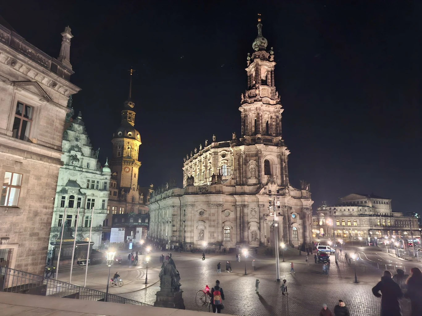 Oświetlony plac miejski otoczony zabytkowymi budynkami, w centrum monumentalna katedra z wysoką wieżą, w tle wieża ratuszowa, przez plac przechadzają się ludzie podziwiający architekturę.