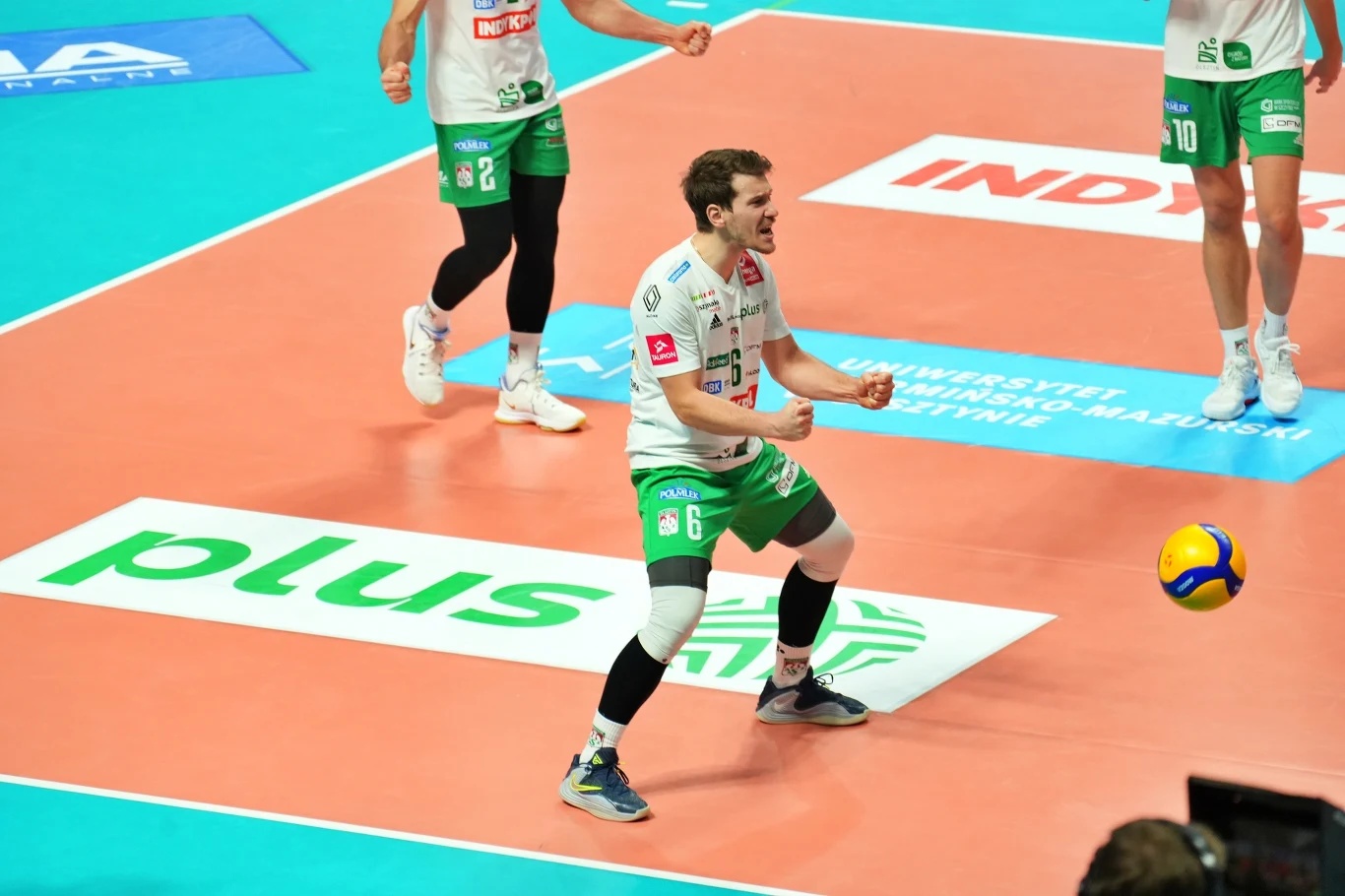 Zawodnik siatkarski w białozielonym stroju energicznie świętuje zdobycie punktu na boisku do siatkówki, wokół niego kilku innych graczy, a na parkiecie widoczna piłka i oznaczenia sponsorskie.