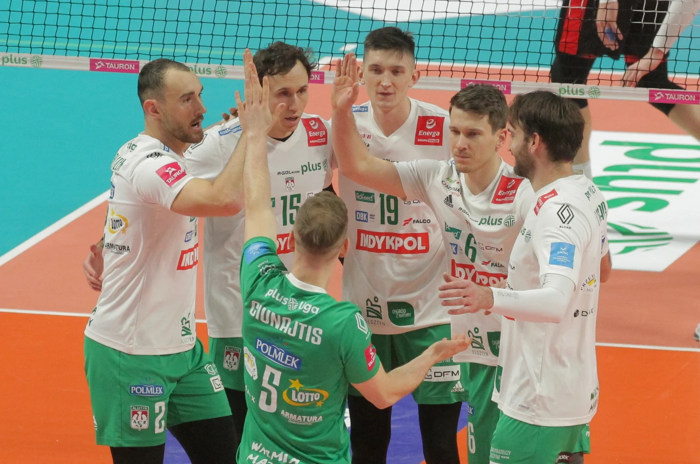 Grupa siatkarzy drużyny Indykpol AZS Olsztyn podczas meczu na parkiecie, zawodnicy ubrani w białe i zielone stroje sportowe, wykonują gest przybijania piątek.