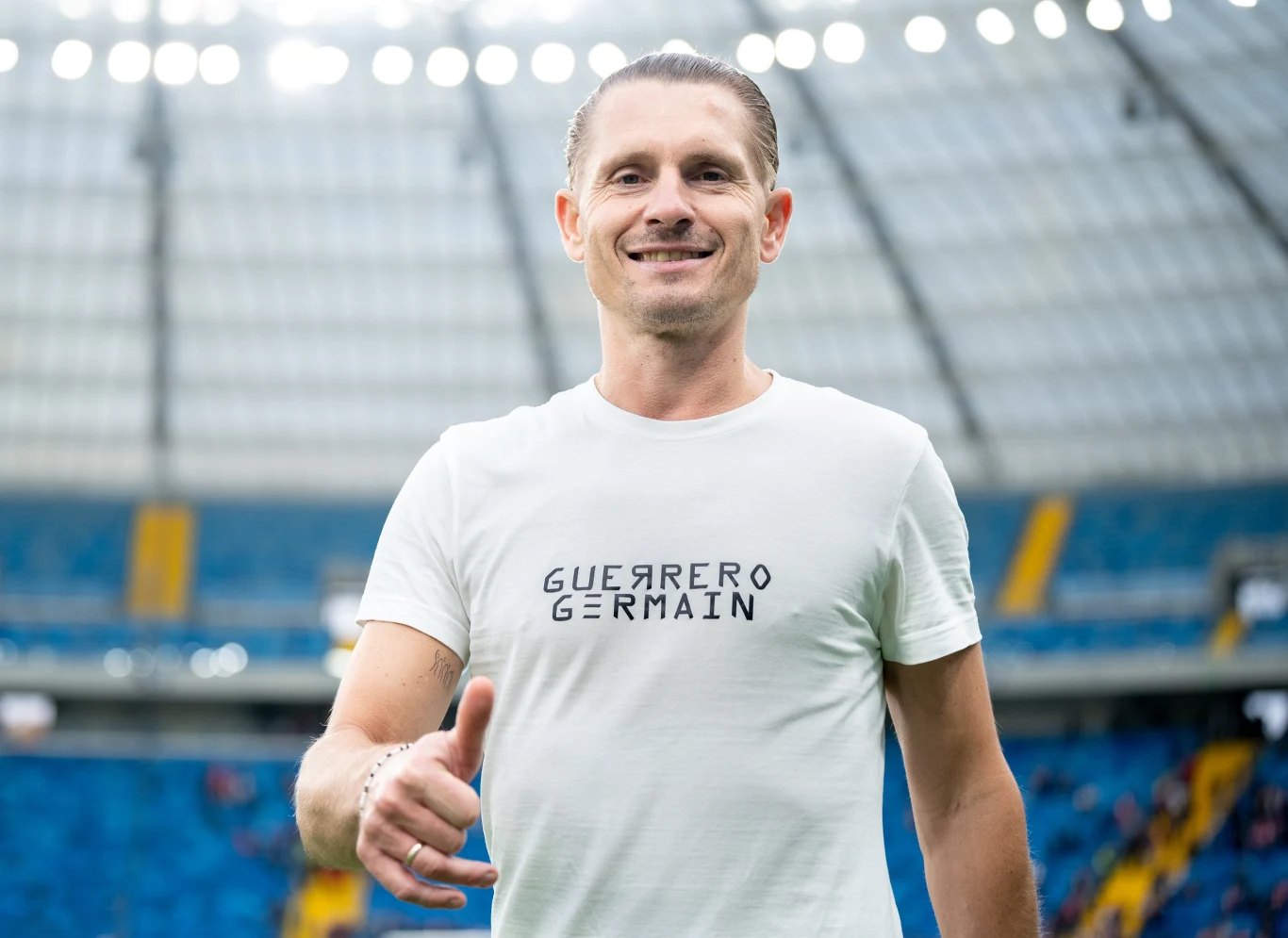 Artur Wichniarek Uśmiechnięty mężczyzna w białej koszulce z napisem GUERRERO GERMAIN na tle stadionu sportowego z niebieskimi trybunami.