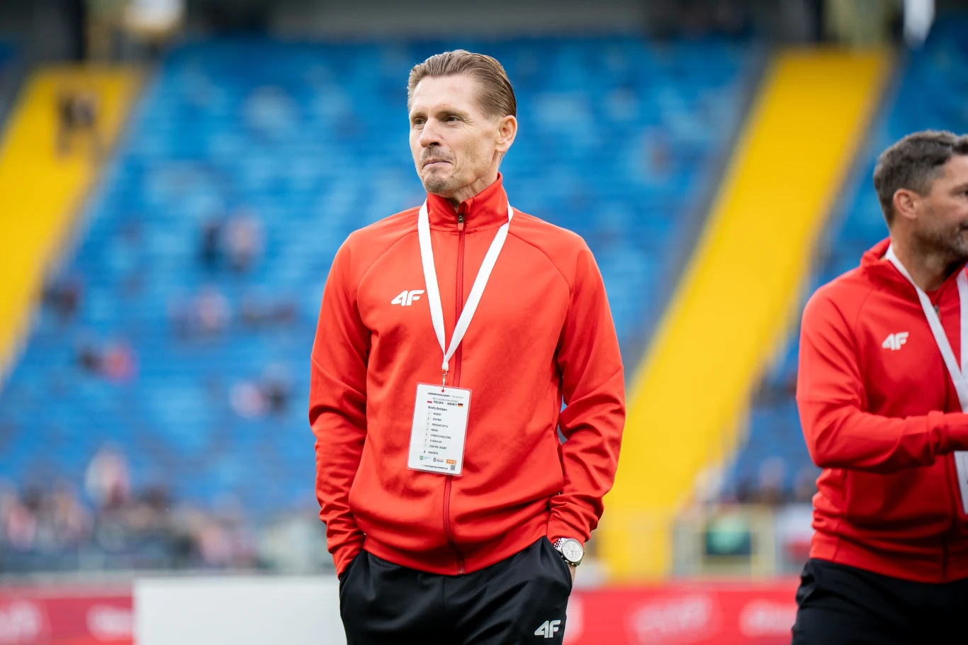 Artur Wichniarek Mężczyzna w czerwonej sportowej kurtce ze smyczą i identyfikatorem na szyi stoi na tle niebieskich trybun stadionu, obok niego inna osoba w podobnym stroju.