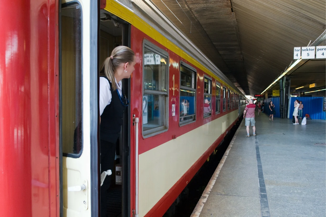 Pamiętasz te wagony PKP? Teraz wracają w jubileuszowym roku Pociąg pasażerski przy peronie na dworcu, konduktorka wygląda z drzwi wagonu, na peronie kilka osób, konstrukcja dachu i tablice z numerami peronów na górze.