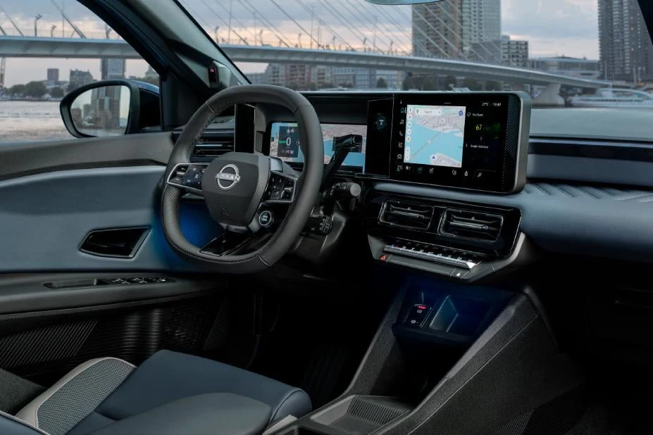 Nowoczesne wnętrze samochodu z cyfrowym kokpitem, dużym ekranem nawigacji, wielofunkcyjną kierownicą z logo Nissan oraz widokiem na miasto przez boczną szybę.