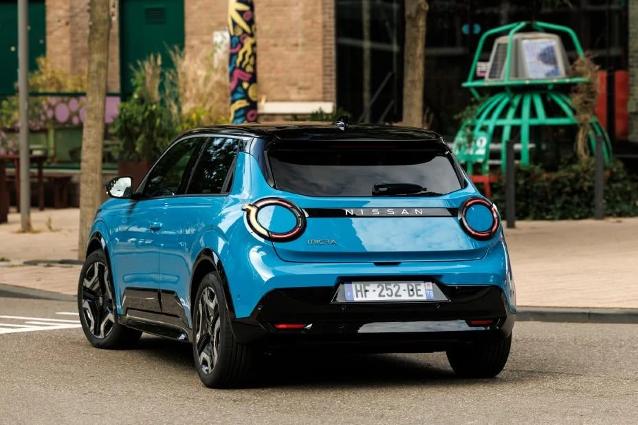 Niebieski samochód Nissan o nowoczesnej sylwetce stoi zaparkowany przy ulicy w otoczeniu miejskiej zabudowy, w tle widoczne drzewa, fragmenty murali i zielona metalowa konstrukcja.