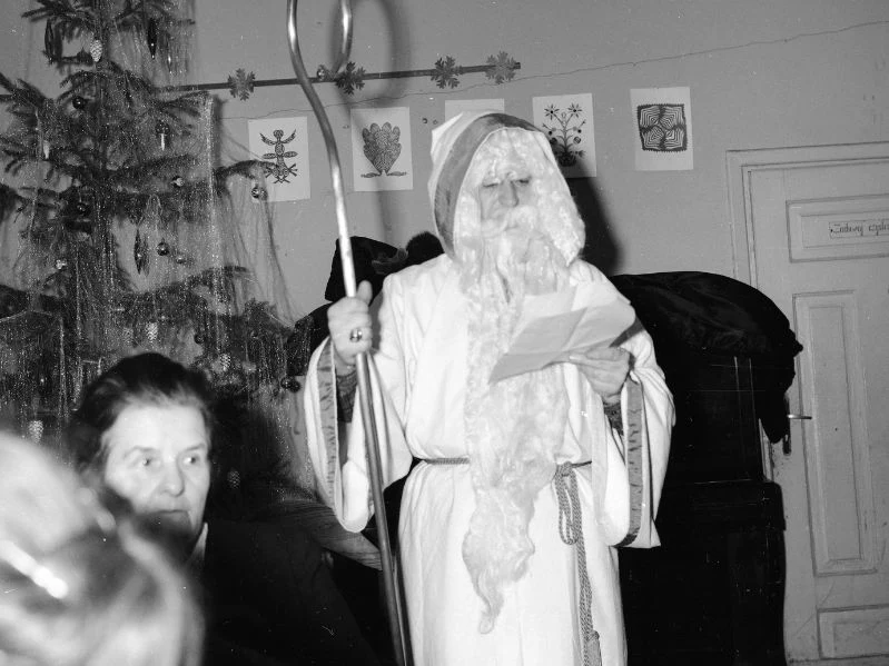 Starszy mężczyzna przebrany w strój Świętego Mikołaja z długą brodą i pastorałem trzyma kartkę, obok widoczna jest choinka i dekoracje ścienne z rysunkami, w pierwszym planie kobieta.
