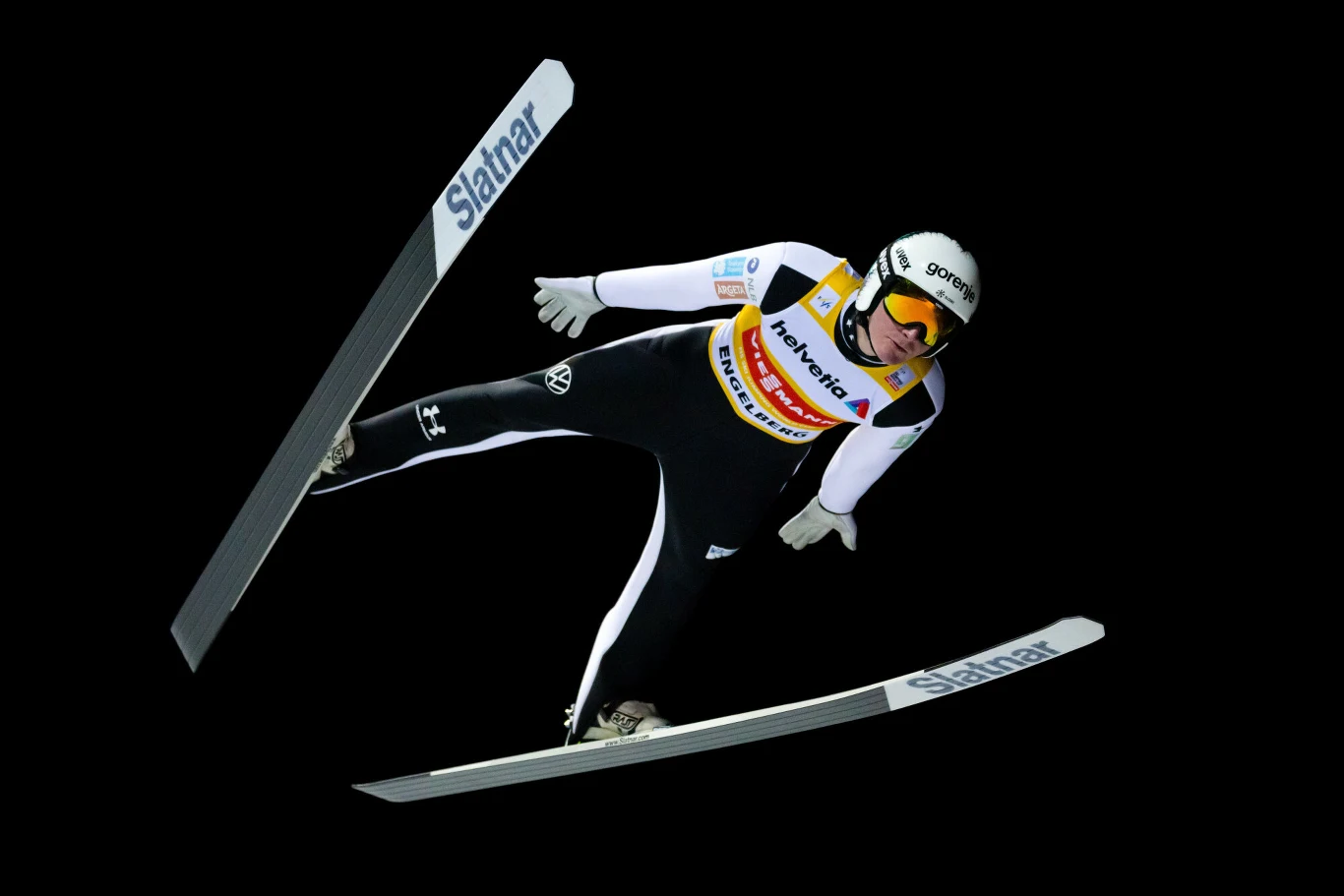 Skoczek narciarski w locie, ubrany w profesjonalny kombinezon i kask, z widocznymi nartami oraz numerem startowym, na czarnym tle podkreślającym sylwetkę sportowca.