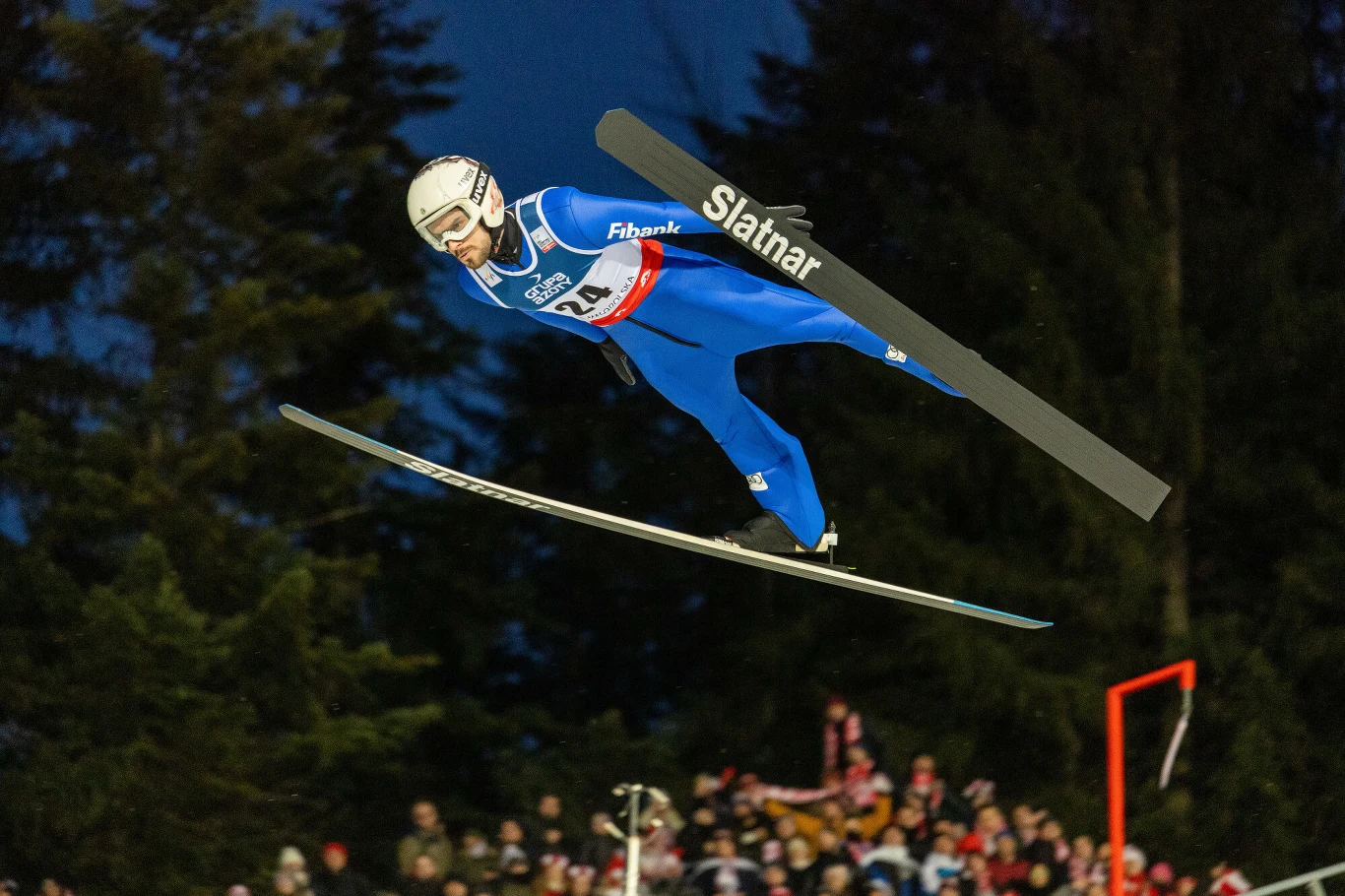 Zawodnik w niebieskim kombinezonie podczas skoku narciarskiego w powietrzu, w tle ciemne drzewa oraz kibice zgromadzeni wzdłuż skoczni.