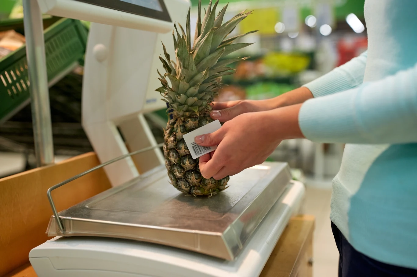 Osoba waży ananasa na elektronicznej wadze sklepowej, trzymając kartę produktową, w tle widoczne warzywa i owoce oraz półki sklepu spożywczego.