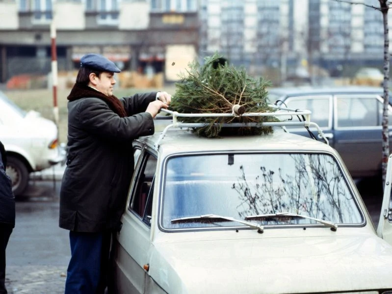 Mężczyzna w zimowym ubraniu mocuje choinkę na dachu małego samochodu na tle bloków mieszkalnych. Na parkingu widoczne są inne pojazdy, a otoczenie sugeruje osiedle miejskie w okresie zimowym.