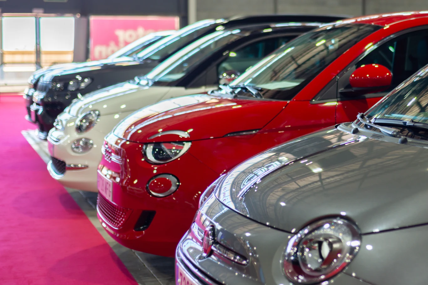 Szereg kolorowych samochodów osobowych ustawionych równolegle na wystawie lub w salonie samochodowym. Lśniące karoserie i wyraźne reflektory skupiają uwagę na detalach motoryzacyjnych.