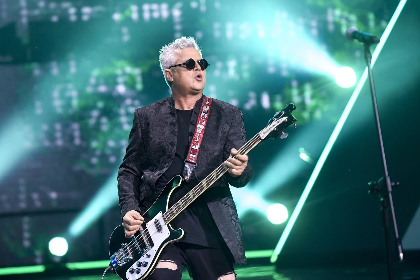 Jacek "Dżej Dżej" Jędrzejak jest muzycznym liderem grupy Big Cyc Muzyk w ciemnych okularach grający na gitarze basowej podczas koncertu na scenie, dynamiczne oświetlenie w tle oraz mikrofon ustawiony z boku przypominają atmosferę muzycznego występu.