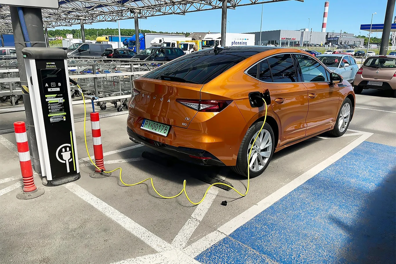 Właściciele elektryków płacą więcej za naprawy i transport auta. Koszty sięgają 20 tys. euro. Pomarańczowy samochód elektryczny przy stacji ładowania na parkingu przed centrum handlowym, otoczony słupkami.