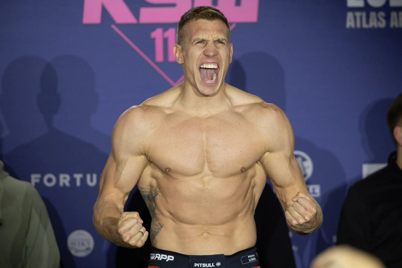 Atletycznie zbudowany mężczyzna bez koszulki wydobywa z siebie okrzyk triumfu, z zaciśniętymi pięściami i napiętymi mięśniami na tle baneru promocyjnego imprezy sportowej. Wokół stoją inne osoby, ale pozostają w cieniu.