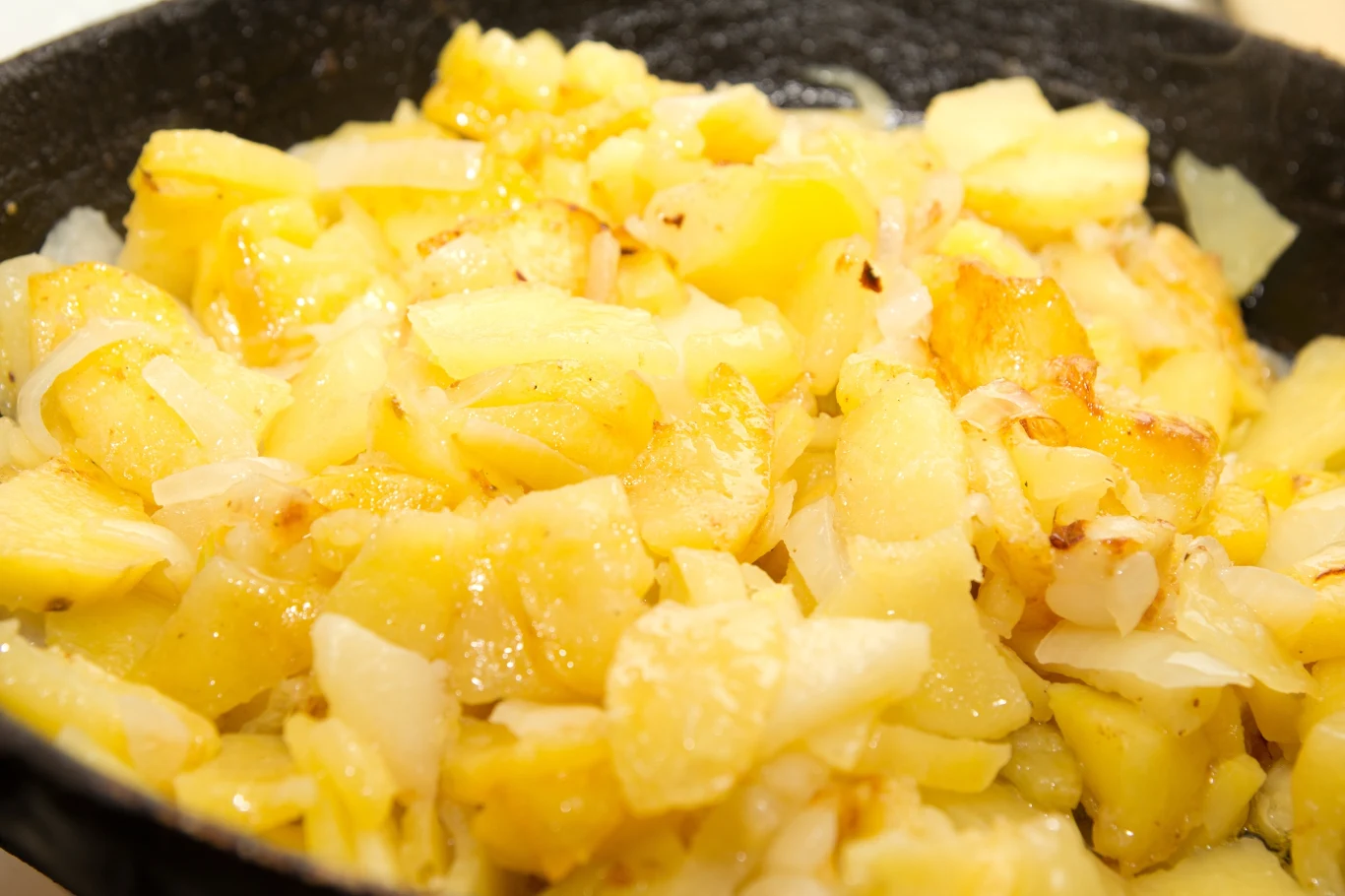 Podsmażone ziemniaki z cebulą na patelni, delikatnie zarumienione i pokrojone w nieregularne kawałki, tworzące apetyczną, złocistą kompozycję.