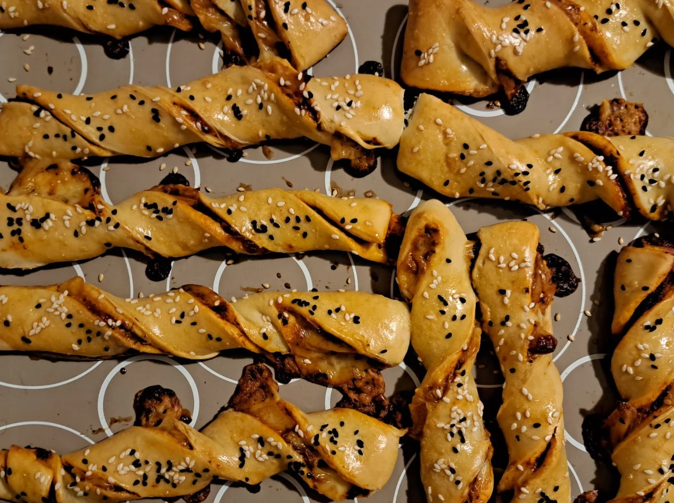 Wypieczone paluchy z ciasta drożdżowego o złocistej skórce, skręcone w spiralę, posypane czarnym i białym sezamem, ułożone w rzędach na papierze do pieczenia z charakterystycznym wzorem.