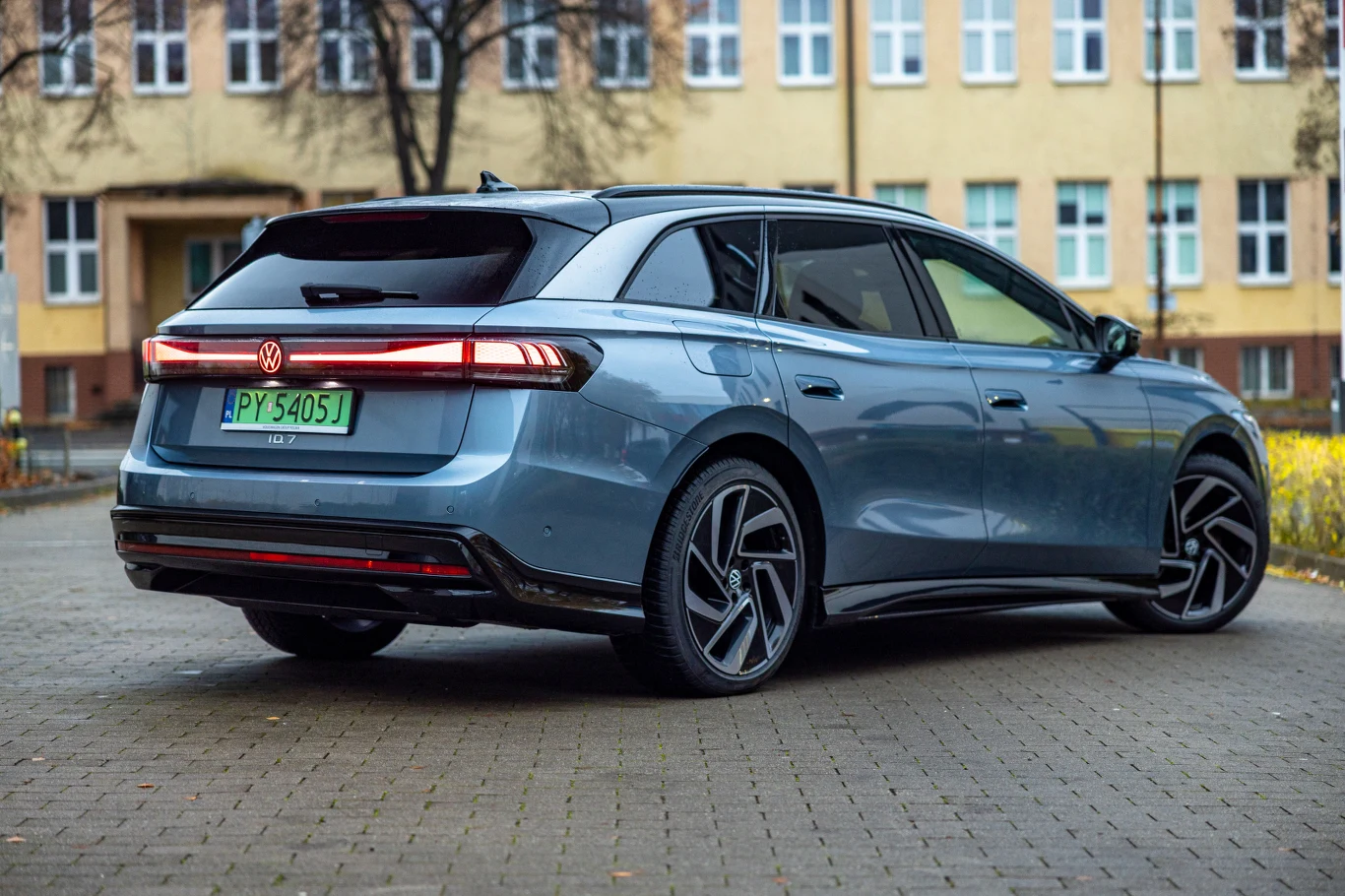ID.7 dostępne jest w wersji Tourer (kombi) oraz jako klasyczny sedan Elektryczny samochód osobowy Volkswagen ID.7 w kolorze niebieskim, zaparkowany na brukowanym placu przed budynkiem z licznymi oknami. Pojazd ma nowoczesny design, zielone tablice rejestracyjne oraz przyciemnione szyby.