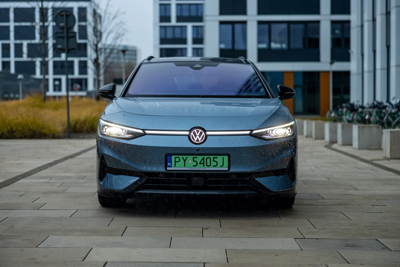 Świecący znaczek i linia łącząca reflektory wpisują się w obecną modę Nowoczesny samochód marki Volkswagen o zielonych tablicach rejestracyjnych stoi na jasnym, brukowanym chodniku otoczonym nowoczesnymi budynkami mieszkalnymi i biurowymi.