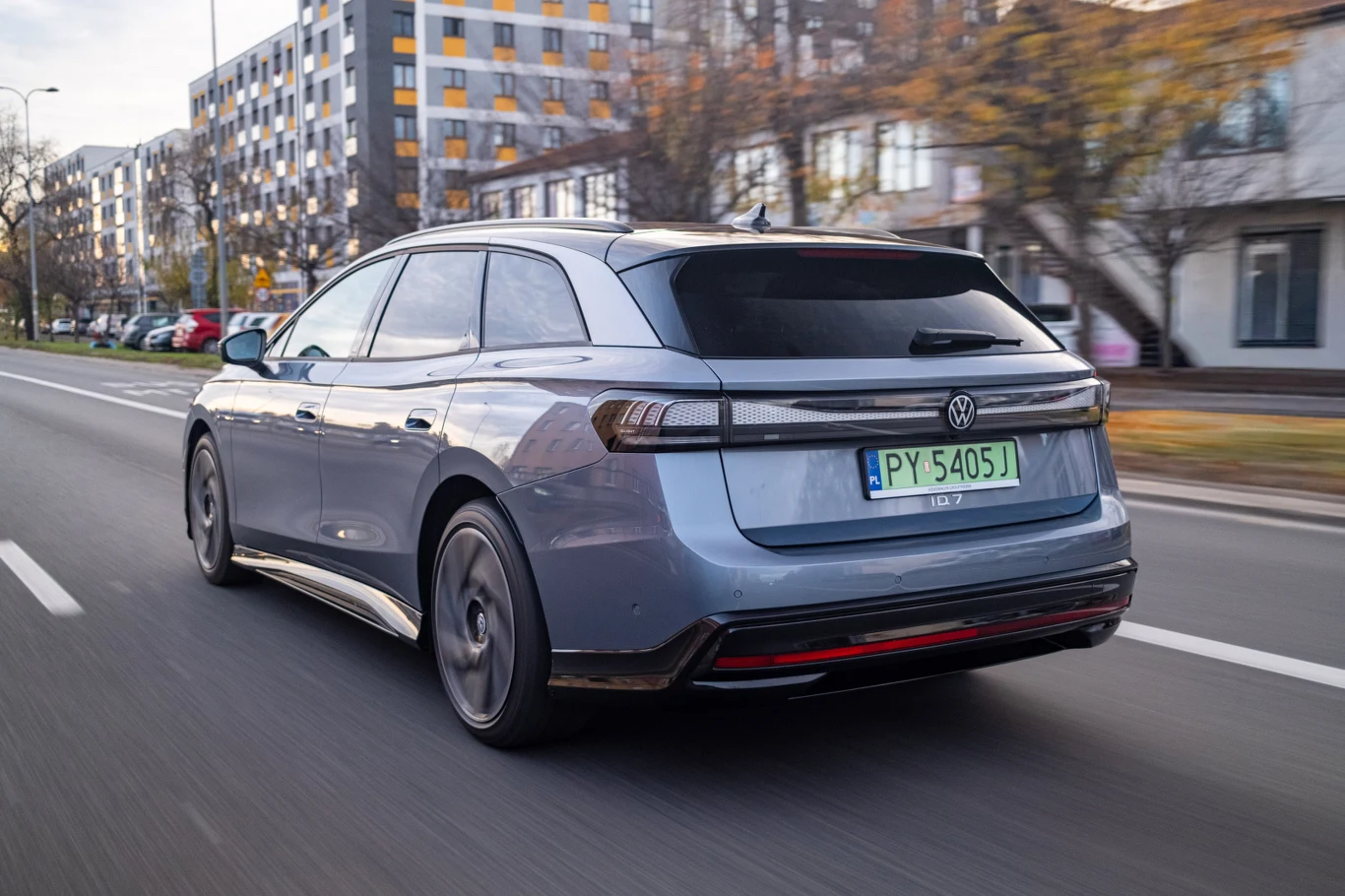 Choć nie zapewnia "atomowych" przyspieszeń, ID.7 bardzo sprawnie przemieszcza się w trasie Szary samochód osobowy marki Volkswagen jedzie miejską ulicą o nowoczesnej zabudowie, widoczne są tablice rejestracyjne oraz rozmyte drzewa i budynki w tle.