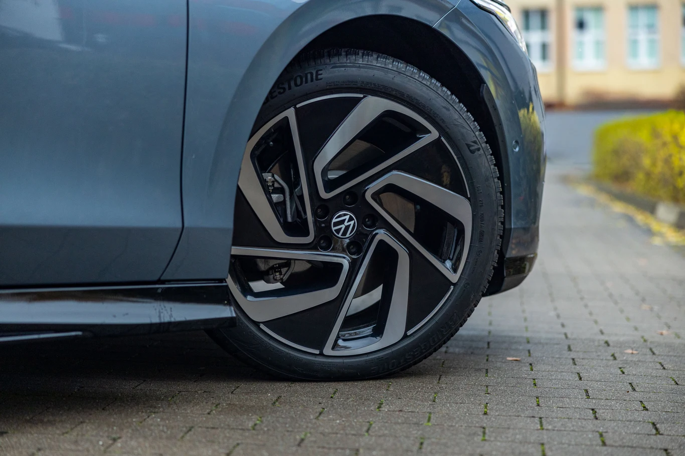 Testowy egzemplarz wyposażono w 20-calowe obręcze. Dostępne są jeszcze o cal większe Nowoczesna felga aluminiowa z wyrazistym wzorem, zamontowana na szarym samochodzie, z łatwo widocznym bieżnikiem opony marki Bridgestone, zaparkowany na kostce brukowej w pobliżu budynku i niskiego żywopłotu.