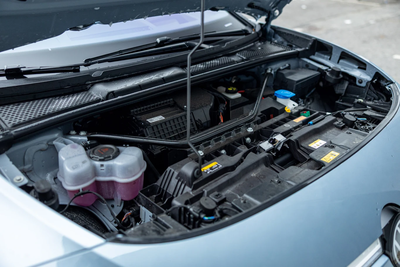 Z przodu Volkswagen nie przewidział żadnego schowka Widok komory silnika samochodowego z otwartą maską, na pierwszym planie zbiornik płynu chłodniczego, podzespoły elektryczne oraz oznaczenia ostrzegawcze.